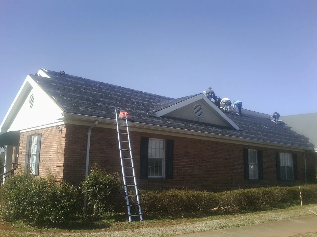 Crew tearing shingles off a brick ranch roof with a ladder set — Emergency Roof Repair work by our crew in Erin