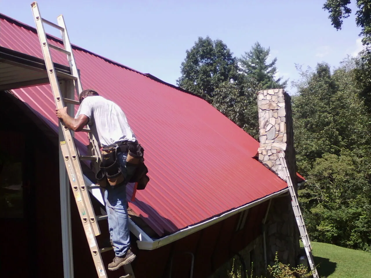 Roofer on a ladder beside a red metal roof and stone chimney — Soffit & Fascia Repair by Roofing Brothers in Erin